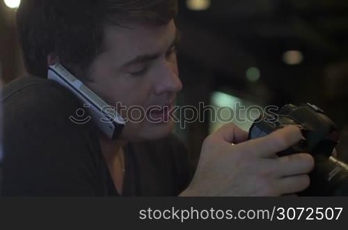 Young man using digital camera while talking on mobile held with shoulder. Discussing the best shots