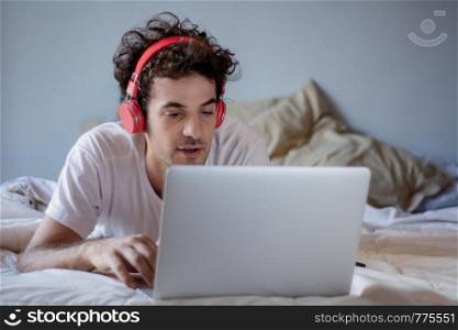 Young man using his laptop with headphones in bed.