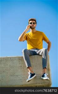 Young man using his smartphone sitting on a ledge outside. Guy wearing sunglasses.. Young man using his smartphone sitting on a ledge outside