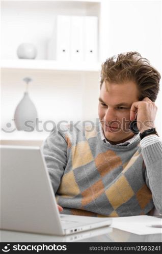 Young man using laptop and calling on phone at home.