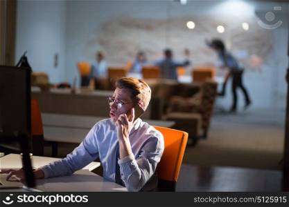 Young man using mobile phone while working on computer at night in dark office.