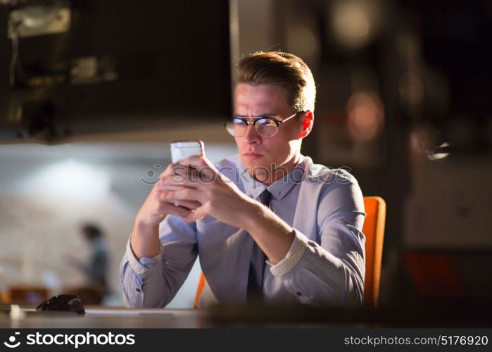 Young man using mobile phone while working on computer at night in dark office.