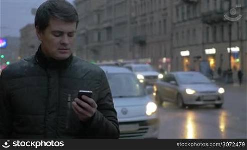 Young man using smart phone in the city on a cold rainy day. He standing by the road with intense traffic in the evening