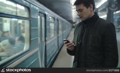Young man using smart phone to type text message at the underground station, train passing by. Everyday traveling by public transport