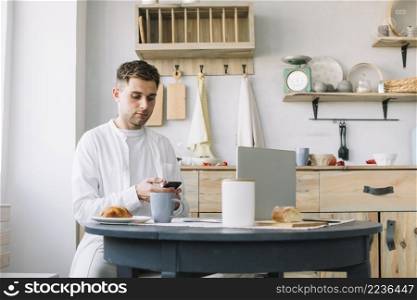 young man using smartphone sitting near table with breakfast laptop kitchen