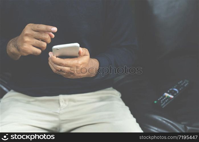 young man using television remote control and mobile smart phone is sitting on a sofa. on or off tv. Leave space to write a description of the message.