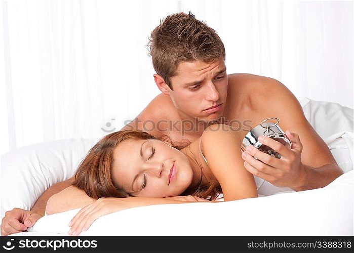 Young man watching alarm clock, woman sleeping in white bed