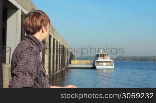 young man watching on river from platform near shore, boat at background in defocus