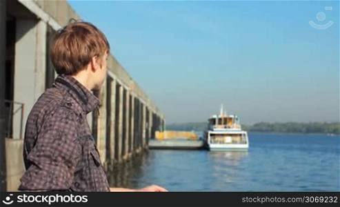 young man watching on river from platform near shore, boat at background in defocus
