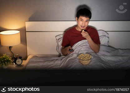 young man watching TV and eating popcorn on a bed at night