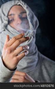 young man wearing a grey hood smoking a cigar with smoke around him against black background