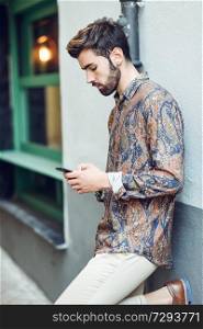 Young man wearing casual clothes looking at his smartphone in the street. Guy with beard and modern hairstyle in urban background. Young man wearing casual clothes looking at his smartphone in the street.