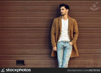 Young man wearing demi-season clothes in the street. Young bearded guy with modern hairstyle with coat, blue jeans and t-shirt against urban wooden blinds.. Young man wearing demi-season clothes in the street.