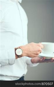 Young man wearing silver wristwatch and white plain shirt holding cup of coffee. Standing in front of plain grey wall