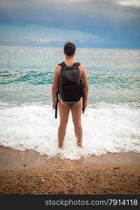 Young man wearing tourist backpack standing on sea shore and looking at horizon