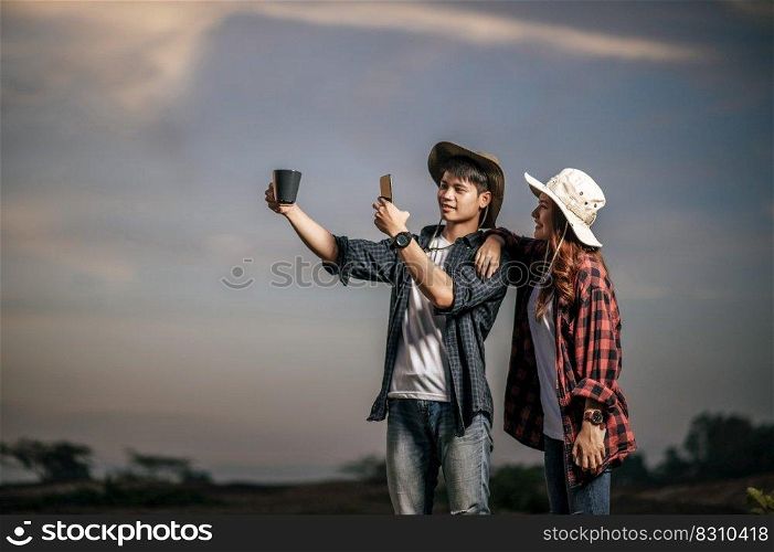 Young man wearing treking hat happy to use smartphone take a photo with coffee mug on beautiful nature in background, Pretty girlfriend standing nearly and smile