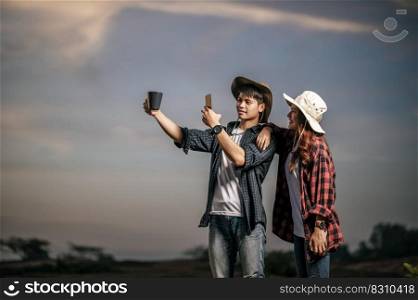 Young man wearing treking hat happy to use smartphone take a photo with coffee mug on beautiful nature in background, Pretty girlfriend standing nearly and smile