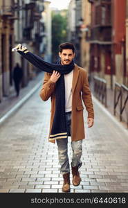 Young man wearing winter clothes in the street. Young bearded guy with modern hairstyle with coat, scarf, blue jeans and t-shirt.