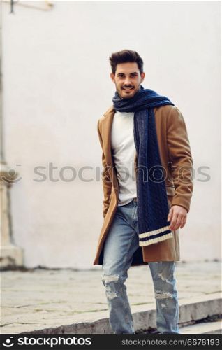 Young man wearing winter clothes in the street. Young bearded guy with modern hairstyle with coat, scarf, blue jeans and t-shirt.. Young man wearing winter clothes in the street.