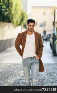 Young man wearing winter clothes in the street. Young bearded guy with modern hairstyle with coat, blue jeans and white t-shirt.. Young man wearing winter clothes in the street. 