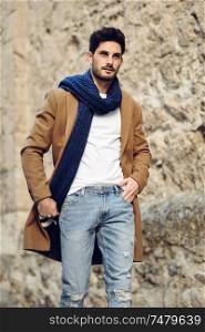 Young man wearing winter clothes in the street. Young bearded guy with modern hairstyle with coat, scarf, blue jeans and t-shirt.. Young man wearing winter clothes in the street.