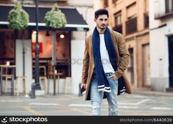 Young man wearing winter clothes with a smartphone in his hand, walking in the street. Young bearded guy with modern hairstyle in urban background.