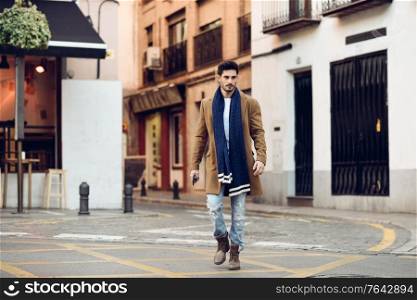 Young man wearing winter clothes with a smartphone in his hand, walking in the street. Young bearded guy with modern hairstyle in urban background.. Young man wearing winter clothes with a smartphone in his hand