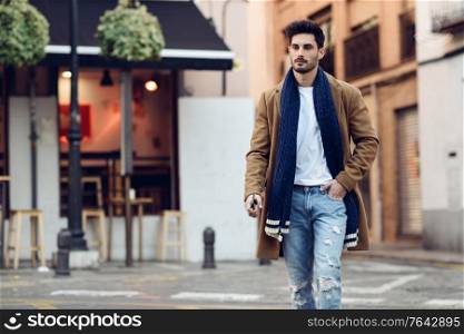 Young man wearing winter clothes with a smartphone in his hand, walking in the street. Young bearded guy with modern hairstyle in urban background.. Young man wearing winter clothes with a smartphone in his hand
