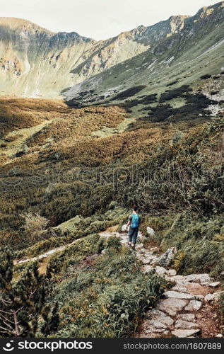 Young man with backpack hiking in a mountains, actively spending summer vacation. Rear view of teenager walking down from top of a hill to valley along mountain footpath. Young man with backpack hiking in a mountains, actively spending summer vacation