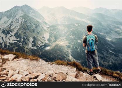 Young man with backpack hiking in a mountains, actively spending summer vacation. Rear view of man standing on the top of a hill admiring mountain landscape panorama. Young man with backpack hiking in a mountains, actively spending summer vacation