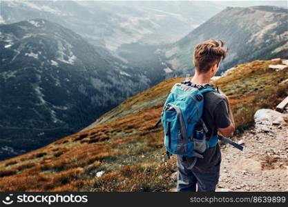 Young man with backpack hiking in a mountains, actively spending summer vacation. Rear view of teenager walking down from top of a hill along mountain path. Young man with backpack hiking in a mountains, actively spending summer vacation