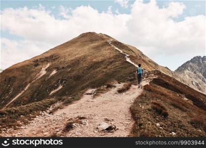 Young man with backpack hiking in a mountains, actively spending summer vacation. Rear view of teenager walking up on a hill along mountain path. Young man with backpack hiking in a mountains, actively spending summer vacation