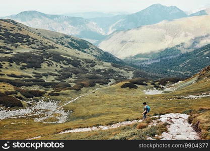 Young man with backpack hiking in a mountains, actively spending summer vacation. Rear view of teenager walking down from top of a hill to valley along mountain footpath. Young man with backpack hiking in a mountains, actively spending summer vacation