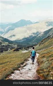 Young man with backpack hiking in a mountains, actively spending summer vacation. Rear view of teenager walking down from top of a hill to valley along mountain footpath. Young man with backpack hiking in a mountains, actively spending summer vacation