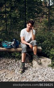 Young man with backpack talking on phone having break sitting on trunk during hike in a mountains actively spending summer vacation time. Young man with backpack talking on phone having break sitting on trunk during hike in a mountains