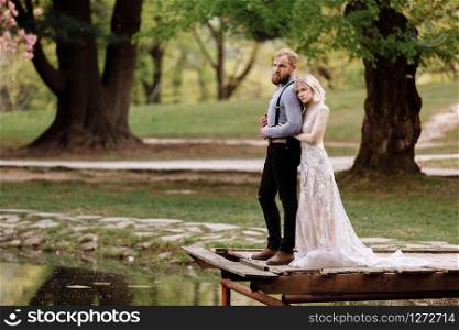 young man with beard and bride in luxury long dress hugging near lake in park with blooming cherry or sakura blossoms on background. Wedding spring day.. young man with beard and bride in luxury long dress hugging near lake in park with blooming cherry or sakura blossoms on background. Wedding spring day