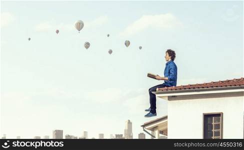 Young man with book. Guy sitting on building roof and reading book