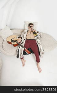 Young man with guitar drinking while lying on sofa