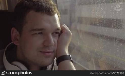 Young man with headphones traveling by rail. He enjoying view from the train window with thoughtful look