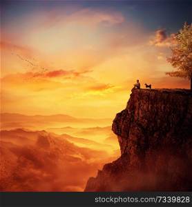 Young man with his faithful dog standing on the peak of a cliff watching the sunset over valley. Recalling childhood memories, friendship between human and animal.