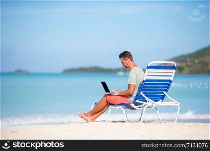Young man with laptop on tropical caribbean beach. Man sitting on the sunbed with computer and working on the beach. Young man with tablet computer during tropical beach vacation