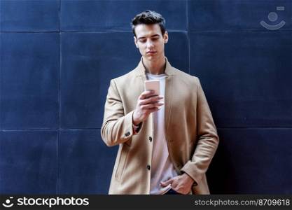 Young man with mobile phone and autumn styling leaning on outdoor wall