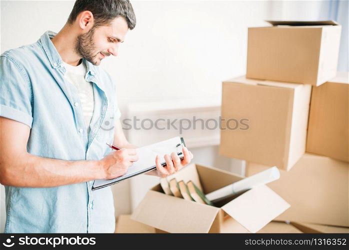Young man with notebook among cardboard boxes, housewarming. Moving to new house