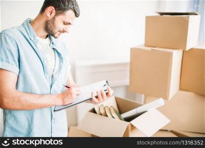 Young man with notebook among cardboard boxes, housewarming. Moving to new house