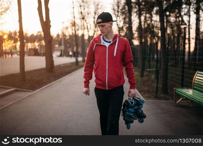 Young man with roller skates in hands walking in city park. Male rollerskater leisure