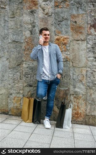 Young man with shopping bags leaning on a stone wall talking on the mobile. Young man with shopping bags and mobile