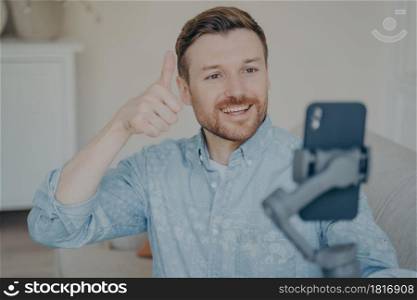Young man with short beard showing thumbs up gesture while video chatting on phone attached to gimbal indoors, talking with his relatives, telling them that everything is fine. Young man showing thumbs up gesture while video chatting on phone