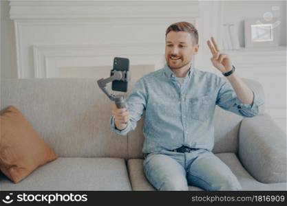 Young man with short beard wearing casual clothes and sitting on comfortable couch, video chatting holding gimbal with phone attached to it, showing peace gesture to camera. Young man showing peace gesture while video chatting