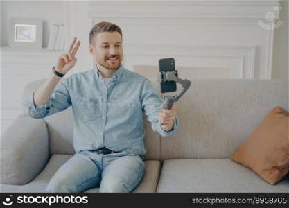 Young man with short beard wearing casual clothes and sitting on comfortable couch, video chatting holding gimbal with phone attached to it, showing peace gesture to camera. Young man showing peace gesture while video chatting