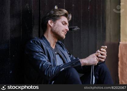 Young man with smartphone earphones sitting in the street black door